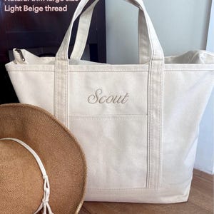 May include: A large off-white canvas tote bag with natural trim and light beige thread embroidery reading "Scout" on the front. The bag features two top handles and a zip closure. A brown woven straw hat with a white cord sits in the foreground.