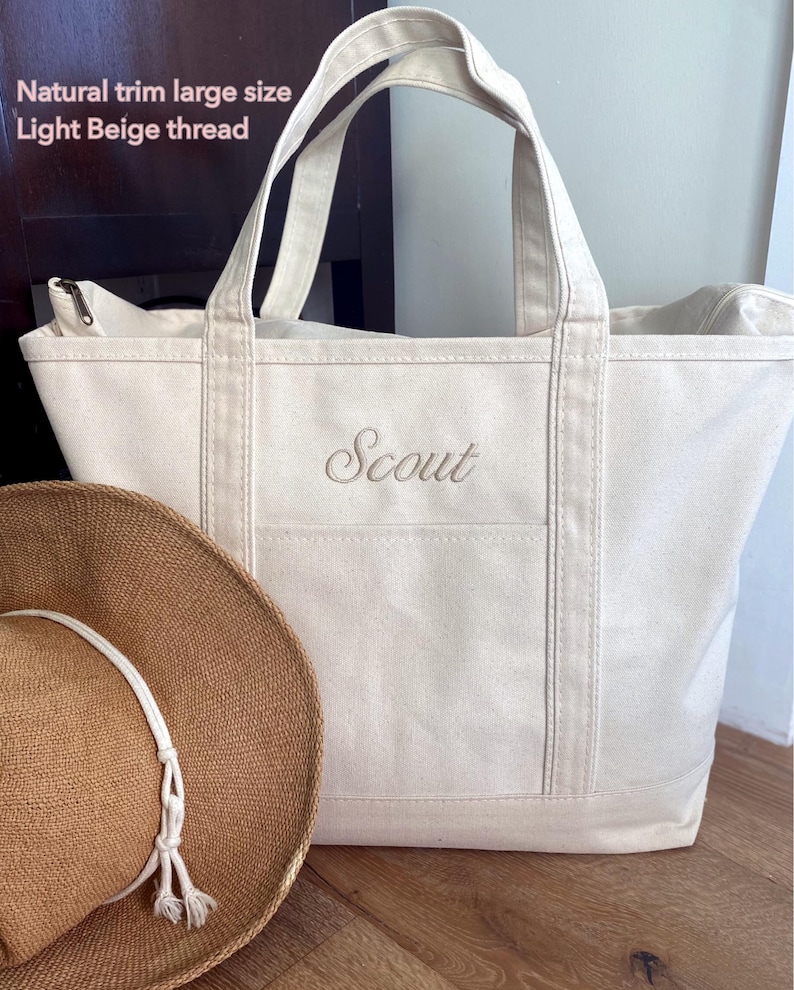 May include: A large off-white canvas tote bag with natural trim and light beige thread embroidery reading "Scout" on the front. The bag features two top handles and a zip closure. A brown woven straw hat with a white cord sits in the foreground.