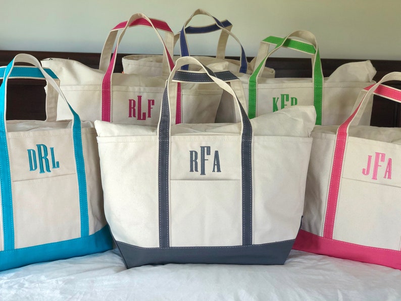 Op de afbeelding: Een collectie off-white canvas draagtassen met gekleurde randen en hengsels. Elke tas is voorzien van een monogram van drie letters, geborduurd in een bijpassende kleur garen, met accenten in turquoise, felroze, marineblauw, limoengroen en donkergrijs.