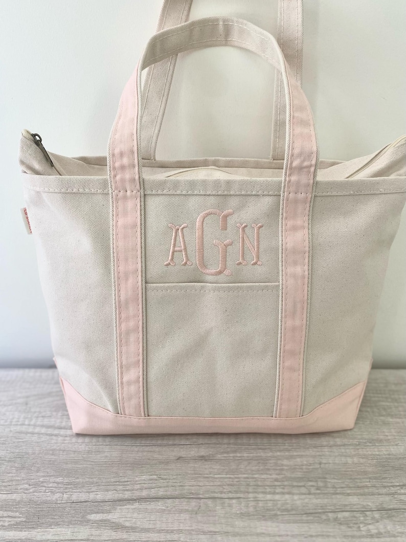 May include: A cream-coloured canvas tote bag featuring a zip closure, two top handles, and a reinforced base in light pink. The front of the bag displays an embroidered monogram with the letters AGN in a light pink thread centred on the panel.