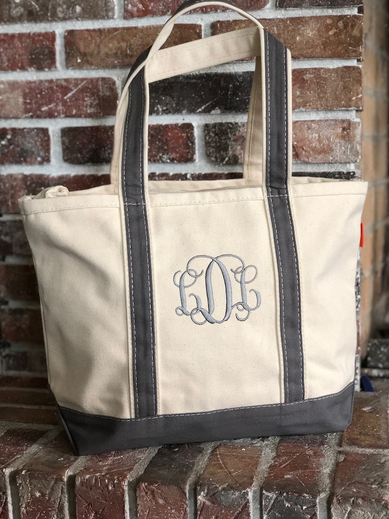 May include: A cream-coloured canvas tote bag featuring dark grey trim along the base and handles. The front of the bag displays a large, embroidered monogram in dark grey thread, consisting of three stylised, interlocking cursive letters.