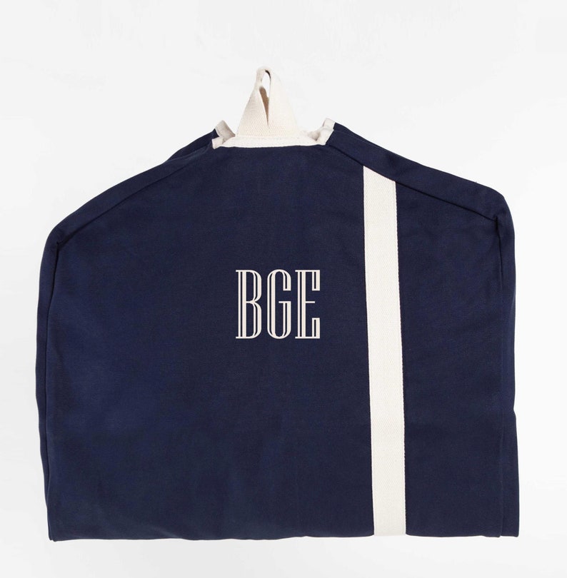 May include: A navy blue garment bag with a white stripe down the side and the monogram "BGE" embroidered on the front.