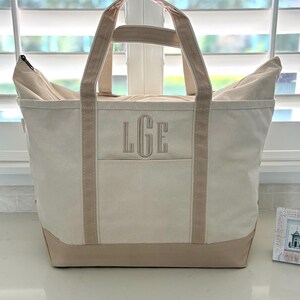 Op de afbeelding: Een cr&egrave;mekleurige canvas draagtas met twee stevige handvatten en een verstevigde onderkant in een beige tint. De voorzijde van de tas toont de geborduurde monogramletters LGE in bijpassend beige garen, gecentreerd boven een horizontale zaknaad.