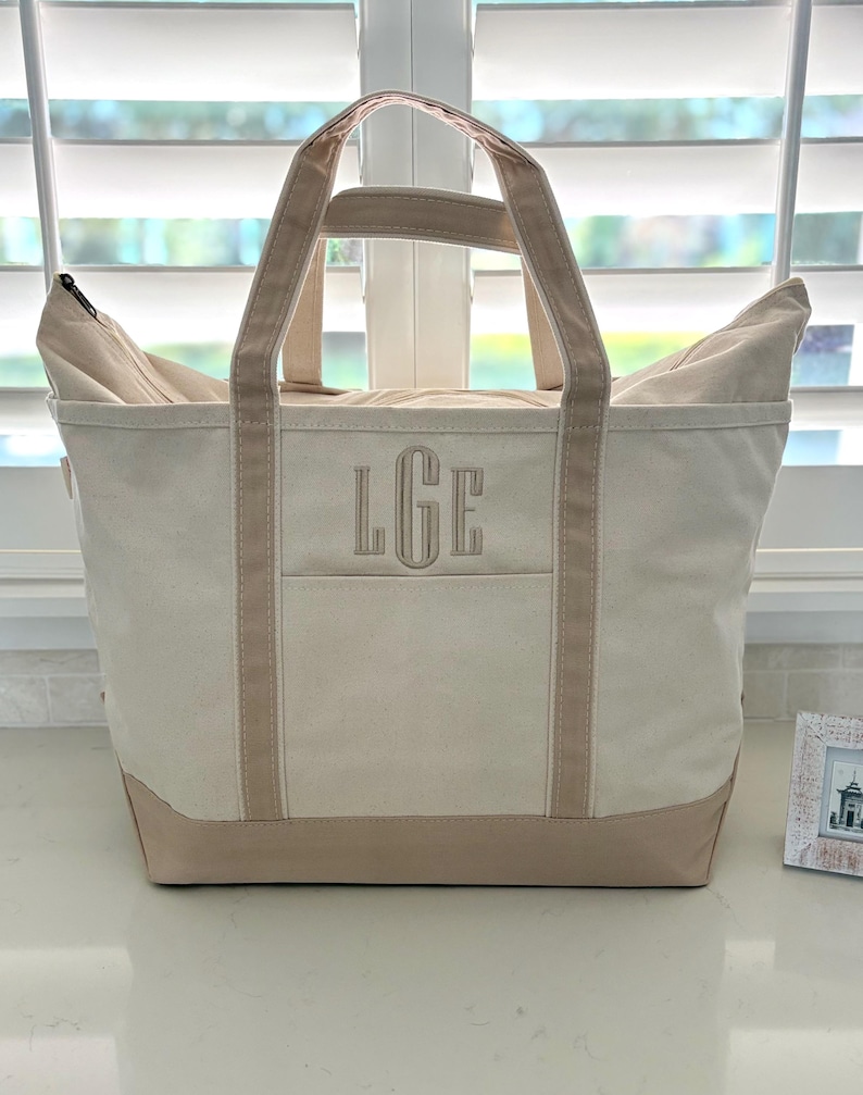 Op de afbeelding: Een cr&egrave;mekleurige canvas draagtas met twee stevige handvatten en een verstevigde onderkant in een beige tint. De voorzijde van de tas toont de geborduurde monogramletters LGE in bijpassend beige garen, gecentreerd boven een horizontale zaknaad.
