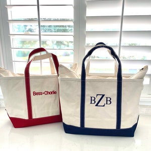 Op de afbeelding: Twee off-white canvas draagtassen met stevige hengsels. De kleinere tas heeft een rode rand en de rode geborduurde tekst "Bess+Charlie". De grotere tas heeft een donkerblauwe rand en het donkerblauwe geborduurde monogram "BZB" op de voorzijde.