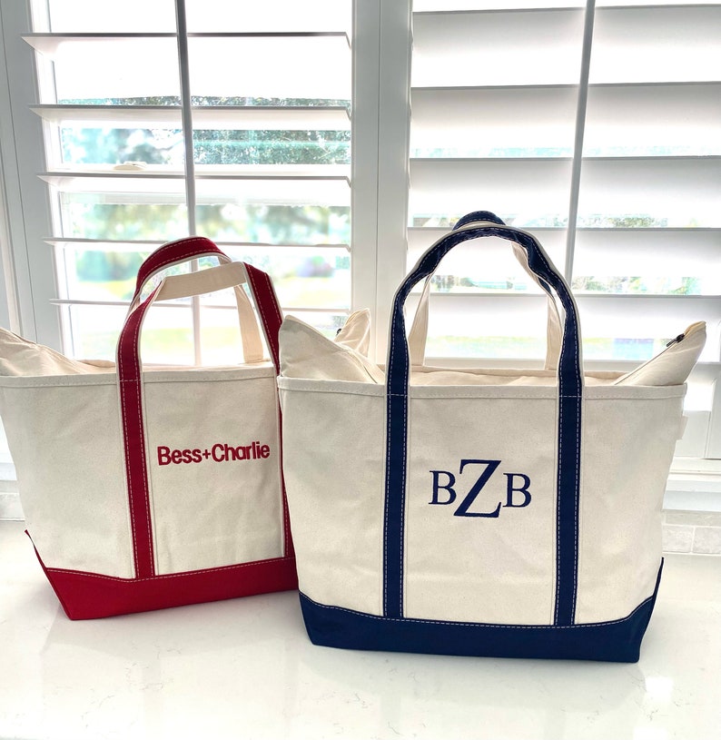 Op de afbeelding: Twee off-white canvas draagtassen met stevige hengsels. De kleinere tas heeft een rode rand en de rode geborduurde tekst "Bess+Charlie". De grotere tas heeft een donkerblauwe rand en het donkerblauwe geborduurde monogram "BZB" op de voorzijde.