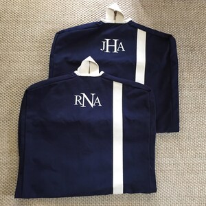 May include: Two navy blue garment bags with white piping and white embroidered initials. The bag on the top has the initials "JHA" and the bag on the bottom has the initials "RNA".