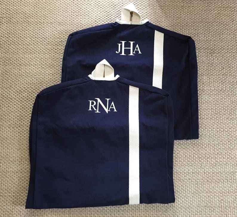 May include: Two navy blue garment bags with white piping and white embroidered initials. The bag on the top has the initials "JHA" and the bag on the bottom has the initials "RNA".