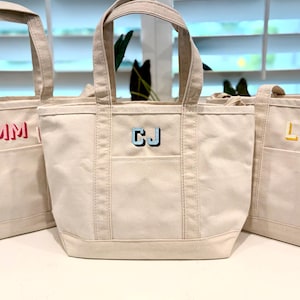 May include: Three canvas tote bags in a neutral color, each with a different set of embroidered initials. The initials are in red, blue, and yellow. Each bag has a front pocket and sturdy handles.