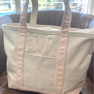 May include: A cream-coloured canvas tote bag featuring light pink trim along the handles and base. The front of the bag displays an embroidered monogram with the letters BMA in a light pink thread, centred above an open exterior pocket.
