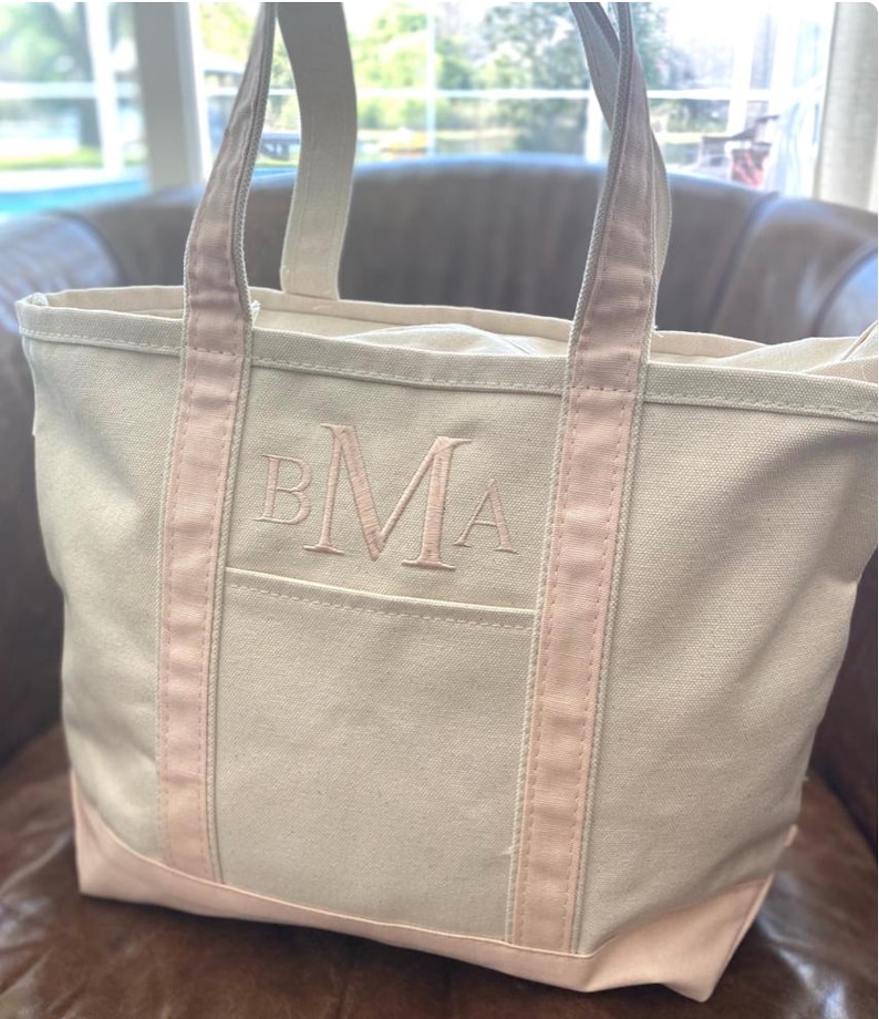 May include: A cream-coloured canvas tote bag featuring light pink trim along the handles and base. The front of the bag displays an embroidered monogram with the letters BMA in a light pink thread, centred above an open exterior pocket.