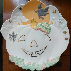 May include: A white ceramic Halloween-themed serving plate shaped like a pumpkin. It features a jack-o'-lantern face, a witch flying in front of a moon, a spiderweb, stars, and a ghost. The plate has gold and pastel accents.