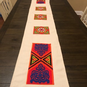 May include: A white table runner with colorful embroidered geometric designs. The designs are in shades of red, orange, yellow, blue, green, and black.