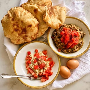 May include: Two white bowls with gold rims containing a savory mixture topped with diced red tomatoes and black pepper. Beside the bowls are three round, toasted pita breads and two whole brown eggs. A silver fork rests inside one of the bowls.