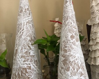 May include: Two white lace cone-shaped Christmas tree decorations. The cones are made of burlap and covered in white lace with a floral pattern. The cones are sitting on a white surface.