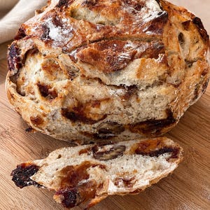 May include: A round loaf of crusty artisan bread with a golden-brown exterior, dusted with flour. The bread is studded with visible dark dried fruit and brown nut pieces throughout the dough, with one slice cut and resting in front of the loaf.