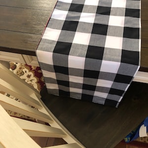May include: Black and white checkered table runner with a red border. The runner is laying on a dark wood table.