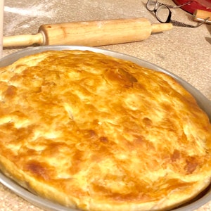 May include: A round, golden-brown baked pastry with a flaky, layered crust sits in a circular metal pan. The surface of the pastry shows variations in color from light tan to deep brown, indicating a crisp texture across the entire top.