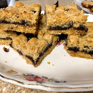 May include: Several square-shaped crumb bars featuring a golden-brown, textured oat-based topping and a thick, dark fruit-based filling layered between the crust, arranged on a white ceramic plate with a delicate floral pattern along the rim.