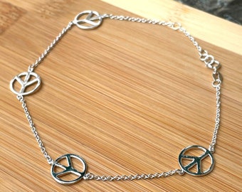 May include: A silver anklet with four peace sign charms. The anklet is on a wooden surface.