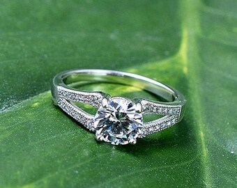 May include: A silver engagement ring with a large, round, clear diamond set in a split shank design. The ring is set on a green leaf.