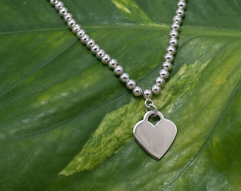 May include: A silver necklace with a heart-shaped pendant. The necklace is made of small, round beads and the pendant is smooth and shiny.