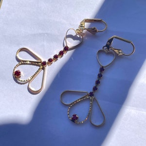 May include: A pair of gold earrings with dangling heart and teardrop shapes. The earrings are adorned with red rhinestones.
