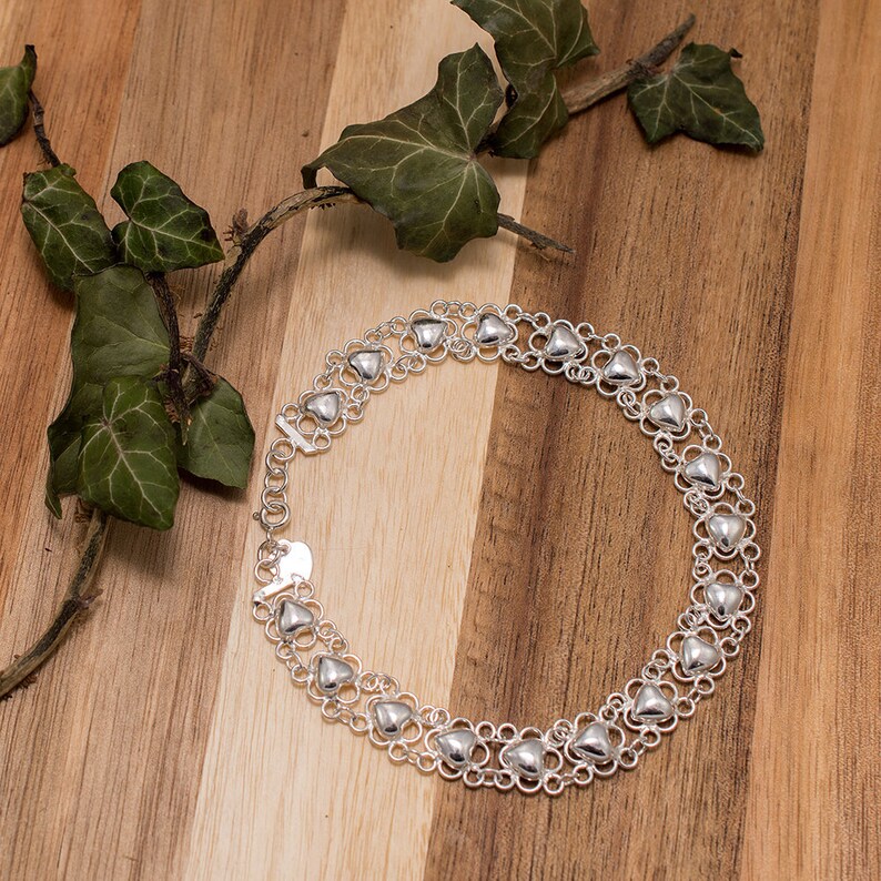 May include: A silver necklace with a double row of heart-shaped links. The necklace is on a wooden surface with green leaves.