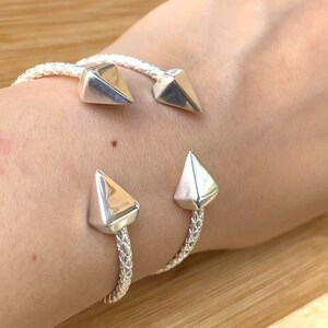 May include: A silver bracelet with two pointed ends. The bracelet is made of a braided metal and has a shiny finish.