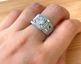 May include: A silver ring with a large, clear, round stone set in a square bezel. The ring has a textured band with a design on one side.