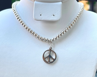 May include: A silver necklace with a peace sign charm. The necklace is made of small, round beads and the charm is a simple, open circle with a peace sign inside.
