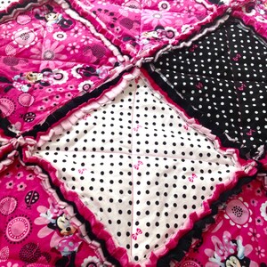 Pode incluir: Uma colcha de retalhos rosa e preta com um centro branco com bolinhas pretas. A colcha &eacute; feita com uma variedade de tecidos, incluindo um tecido rosa com padr&atilde;o floral e um tecido preto com bolinhas brancas. A colcha &eacute; guarnecida com um babado rosa.