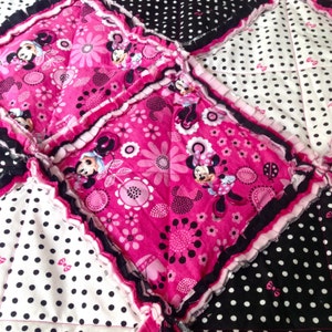 Pode incluir: Uma colcha de retalhos rosa e preta com estampa de Minnie Mouse. A colcha &eacute; feita de quadrados de tecido com diferentes padr&otilde;es, incluindo bolinhas, flores e personagens da Minnie Mouse.