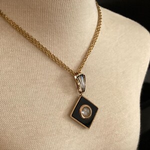 May include: A gold-toned necklace with a black and gold pendant. The pendant is a diamond shape with a black background and a circular, clear stone in the center. The necklace is displayed on a cream-colored mannequin.