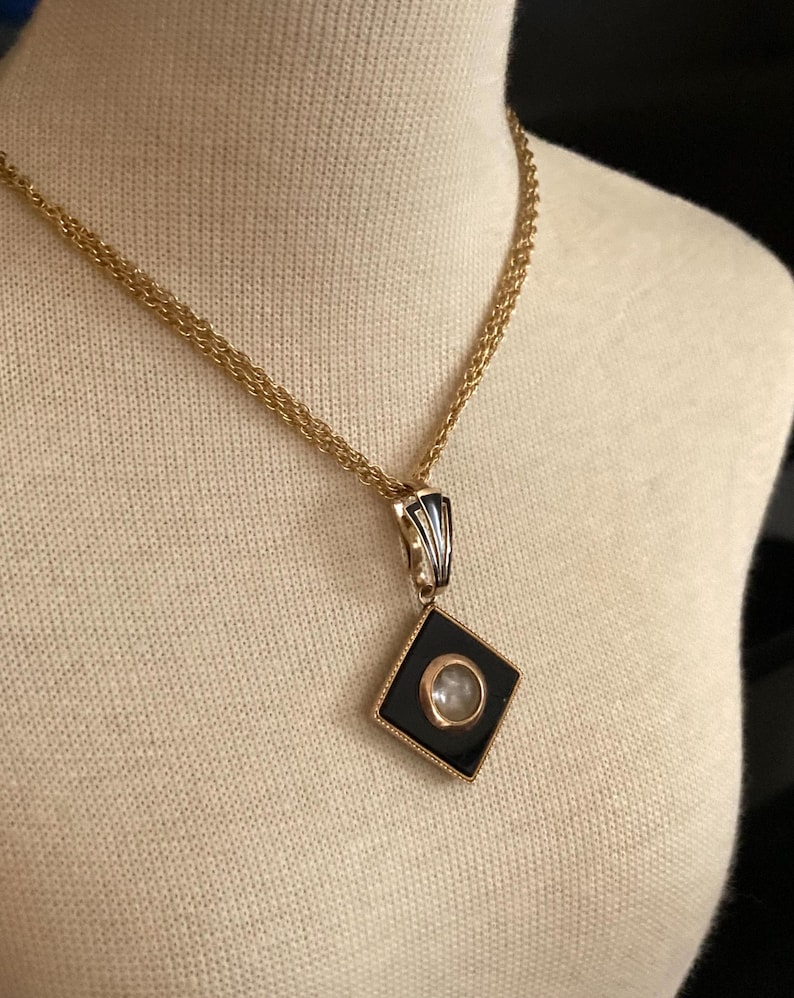 May include: A gold-toned necklace with a black and gold pendant. The pendant is a diamond shape with a black background and a circular, clear stone in the center. The necklace is displayed on a cream-colored mannequin.