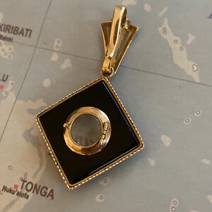 May include: A gold-toned pendant with a black square center and a circular, clear element. The pendant has a decorative bail and a detailed frame. The background is a map.