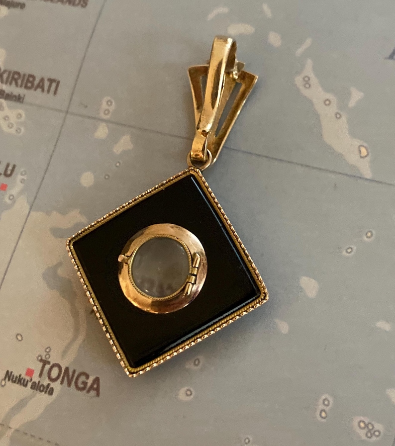 May include: A gold-toned pendant with a black square center and a circular, clear element. The pendant has a decorative bail and a detailed frame. The background is a map.
