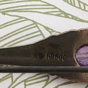 Op de afbeelding: Close-up van een zilveren broche met een paarse steen en de inscriptie "(19)80C".