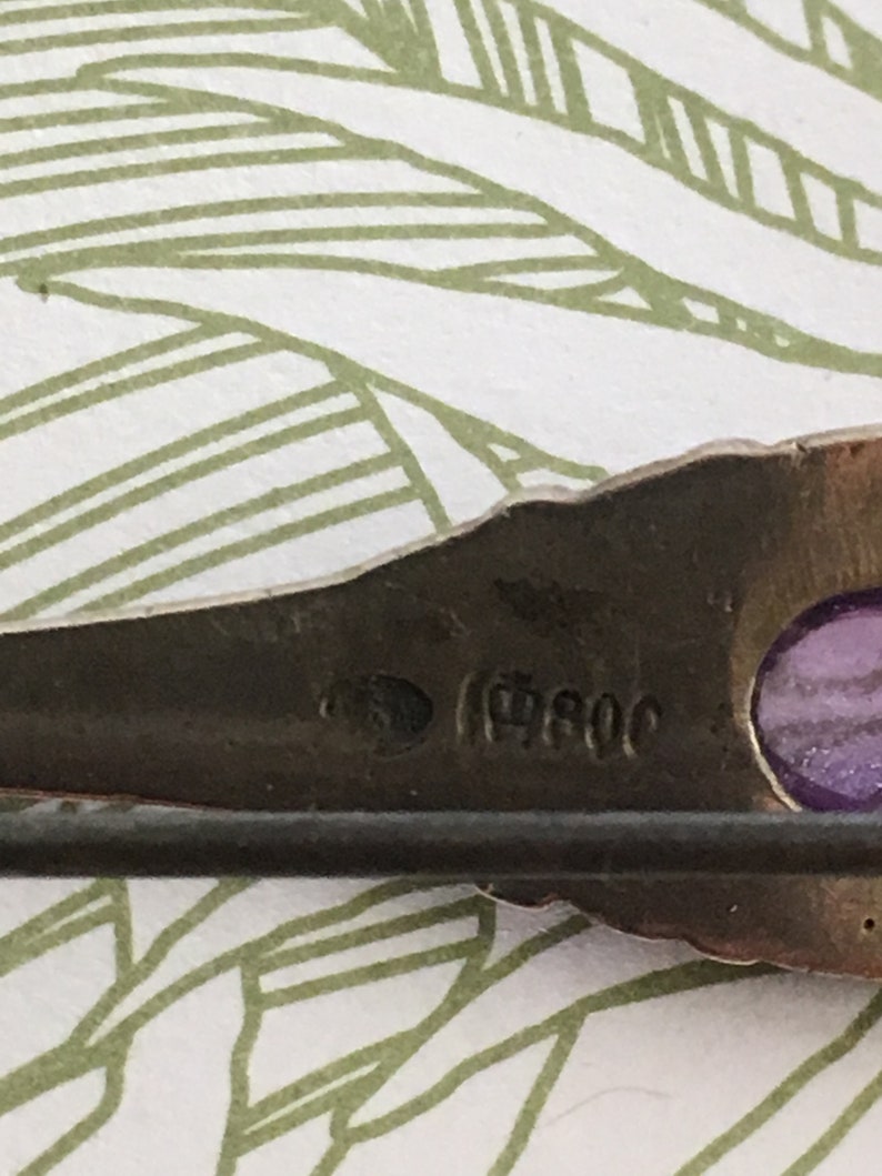 Op de afbeelding: Close-up van een zilveren broche met een paarse steen en de inscriptie "(19)80C".