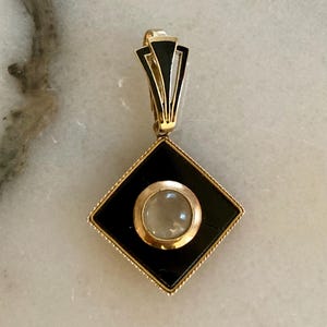 May include: A gold pendant with a black diamond-shaped center and a circular, light-colored stone. The pendant has a gold bail with black accents and a decorative gold border. The design is Art Deco inspired.
