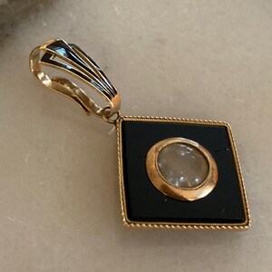 May include: A gold-toned pendant with a black square center and a circular, light-colored inset. The pendant has a decorative bail with black and gold accents. The square frame has a beaded edge.