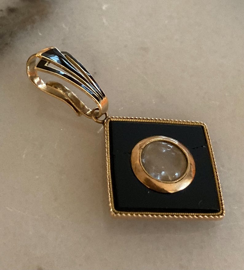 May include: A gold-toned pendant with a black square center and a circular, light-colored inset. The pendant has a decorative bail with black and gold accents. The square frame has a beaded edge.