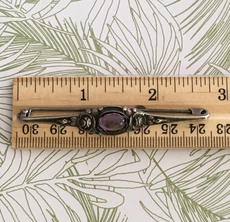 Op de afbeelding: Een zilveren broche met een paarse edelsteen in een bloemmotief. De broche is ongeveer 3,8 cm lang.