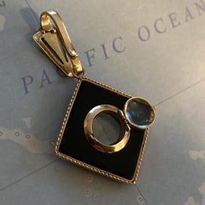 May include: A vintage gold-toned pendant with a black square center, a magnifying glass, and a hinged cover. The pendant has a clip for attachment and is set against a map background.