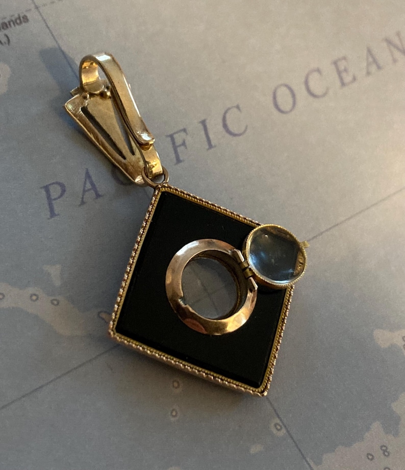 May include: A vintage gold-toned pendant with a black square center, a magnifying glass, and a hinged cover. The pendant has a clip for attachment and is set against a map background.