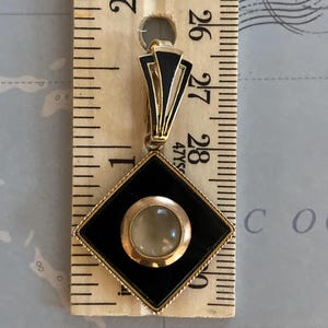 May include: Art Deco pendant with a black and gold geometric design. The pendant features a central, round, light-colored stone set within a gold frame, encased in a black diamond shape. The pendant is approximately 1.25 inches long.
