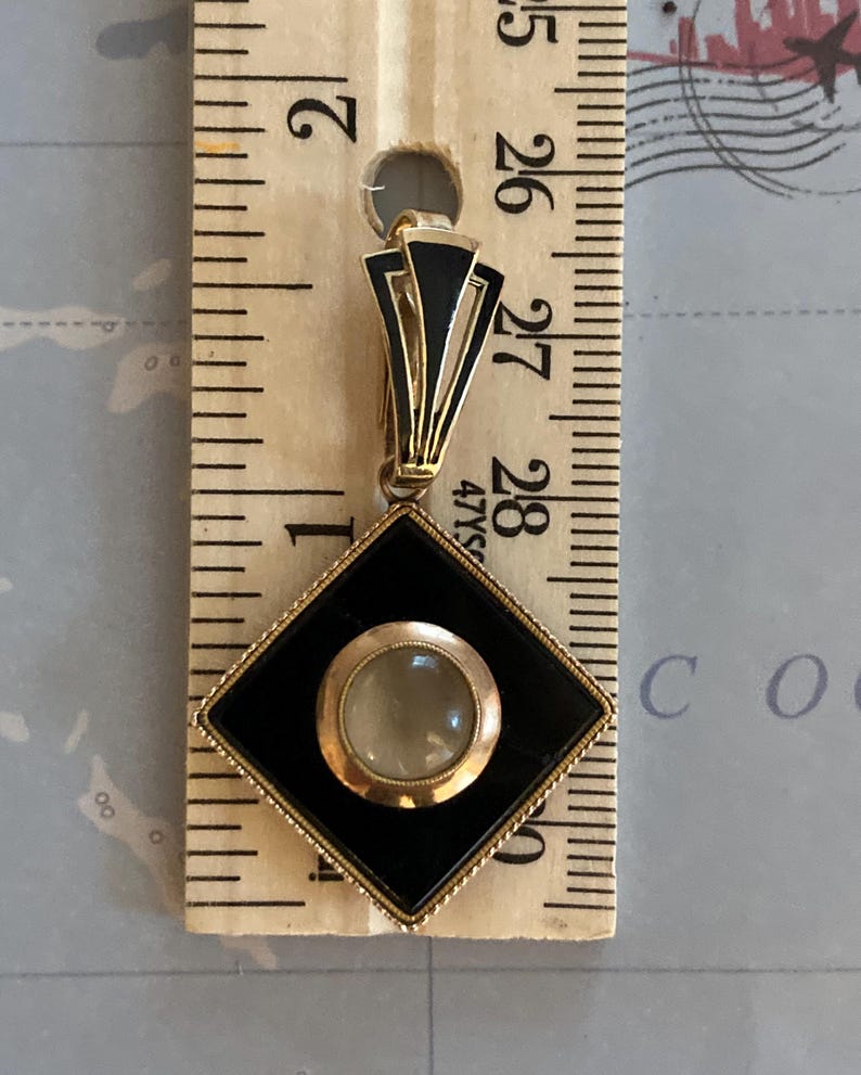 May include: Art Deco pendant with a black and gold geometric design. The pendant features a central, round, light-colored stone set within a gold frame, encased in a black diamond shape. The pendant is approximately 1.25 inches long.