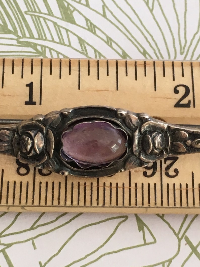 Op de afbeelding: Een zilveren ring met een paarse edelsteen gezet in een bloemmotief. De ring heeft een diameter van ongeveer 2,5 cm.