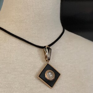 May include: A black and gold pendant necklace with a diamond-shaped pendant. The pendant features a black center with a circular, light-colored stone and gold trim. The necklace has a black cord.