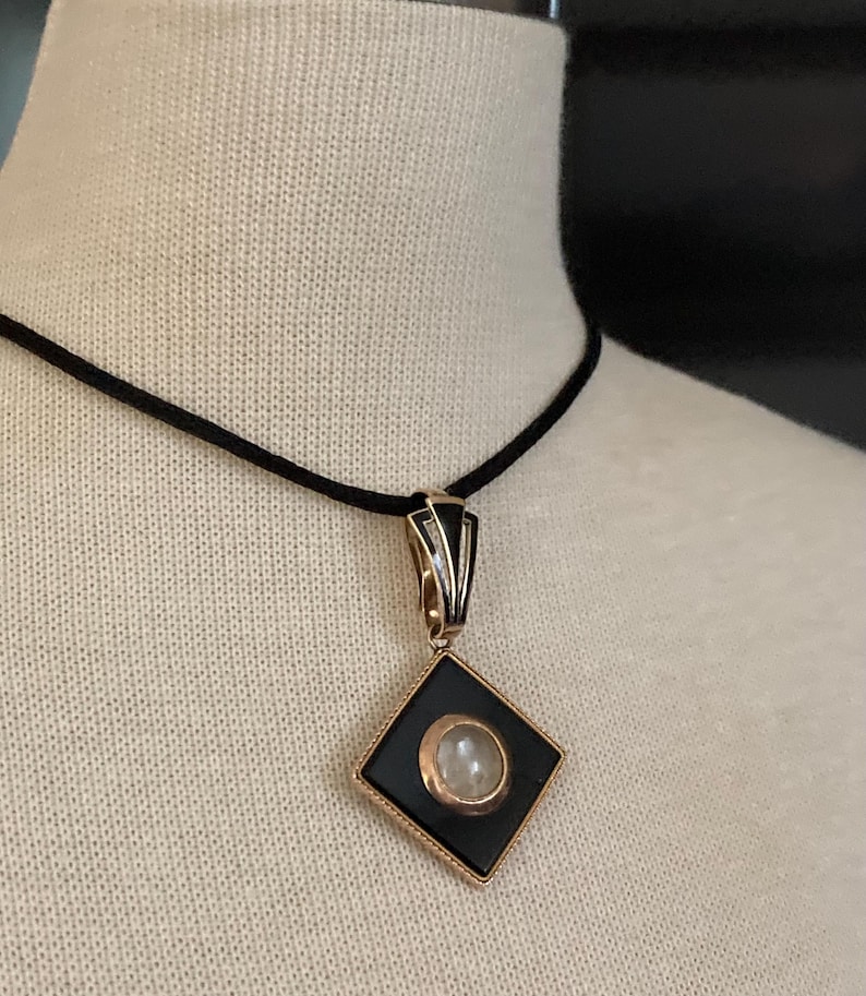 May include: A black and gold pendant necklace with a diamond-shaped pendant. The pendant features a black center with a circular, light-colored stone and gold trim. The necklace has a black cord.